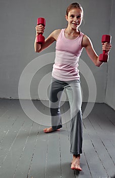 Teenage sportive girl is doing exercises with dumbbells to develop muscles on grey background. Sport healthy lifestyle concept. Sp