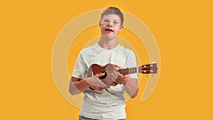 Teenaged disabled boy with Down syndrome having fun while playing ukulele, standing isolated over yellow background