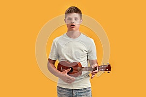 Teenaged disabled boy with Down syndrome looking at camera while playing ukulele, standing isolated over yellow