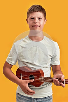 Teenaged disabled boy with Down syndrome looking at camera while playing ukulele, standing isolated over yellow