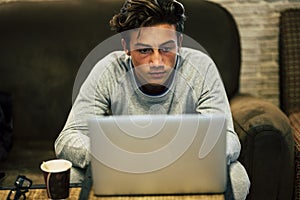 Teenager alone at thome on the sofa with his laptop working or playing or watching videos - night with cofee and glasses on the