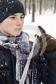 Teenager boy in a blue jacket, in a checkered scarf and in a dark blue hat with a icicle in his hands