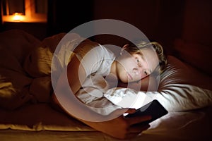 Teenager boy chatting by phone, writing in blog or reading social networks instead sleeping. Young guy laying in bed and watching