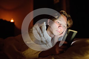 Teenager boy chatting by phone, writing in blog or reading social networks instead sleeping. Young guy laying in bed and watching