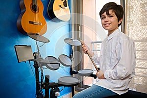 Teenager boy drummer musician in white shirt and blue jeans, sitting at drum set in his retro music studio , holding