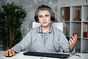 A teenager boy in headphones playing computer games at pc at home in the interior in front of a computer monitor