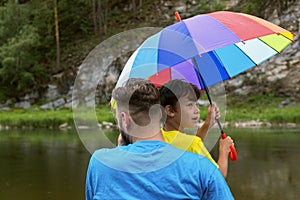 Teenager boy and his father are under colorful umbrella on beach by river or lake