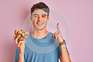 Teenager boy holding bunch of natural walnuts over pink isolated background surprised with an idea or question pointing finger