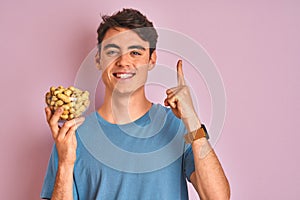 Teenager boy holding a bunch of peanunts over isolated pink background surprised with an idea or question pointing finger with