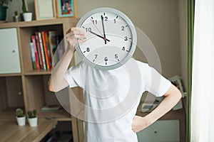 A teenager boy holds a big clock in his hands. A child in a white T-shirt, watch covers his face, a bow on his belt. In the
