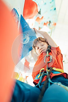 Teenager boy at indoor climbing wall hall. Boy is climbing using a top rope and climbing harness. He hanging on climbing holds and