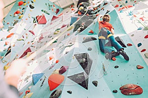 Teenager boy at indoor climbing wall hall. Boy is climbing using a top rope and climbing harness and somebody belaying him from