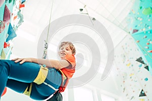 Teenager boy at indoor climbing wall hall. Boy is climbing using a top rope and climbing harness and somebody belaying him from fl photo