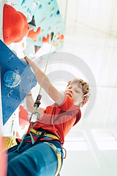Teenager boy at indoor climbing wall hall. Boy is climbing using a top rope and climbing harness and somebody belaying him from fl