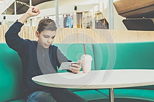 Teenager boy sits at cafe table, plays mobile games on smartphone. Boy is sitting with his hand up, victory, winnings.