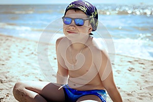 A teenager boy sitting on the sand in swimming trunks, blue sun glasses, is engaged in meditation, relaxation and yoga on the sea