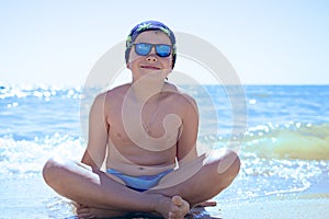 A teenager boy sitting on the sand in swimming trunks, blue sun glasses, is engaged in meditation, relaxation and yoga on the sea