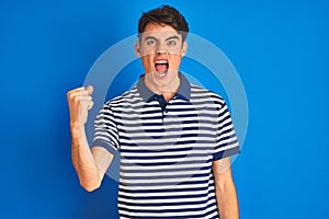Teenager boy wearing casual t-shirt standing over blue  background angry and mad raising fist frustrated and furious while