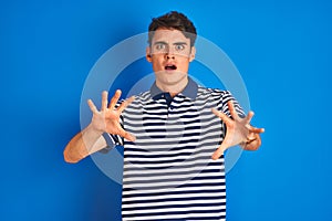 Teenager boy wearing casual t-shirt standing over blue isolated background afraid and terrified with fear expression stop gesture