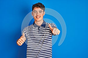 Teenager boy wearing casual t-shirt standing over blue isolated background approving doing positive gesture with hand, thumbs up
