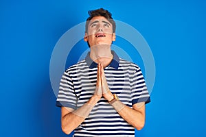 Teenager boy wearing casual t-shirt standing over blue isolated background begging and praying with hands together with hope