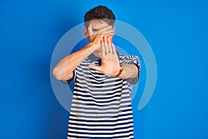 Teenager boy wearing casual t-shirt standing over blue isolated background covering eyes with hands and doing stop gesture with