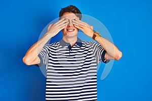Teenager boy wearing casual t-shirt standing over blue isolated background covering eyes with hands smiling cheerful and funny