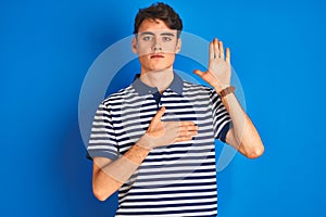 Teenager boy wearing casual t-shirt standing over blue isolated background Swearing with hand on chest and open palm, making a