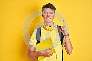 Teenager boy wearing headphones and backpack reading a book over isolated background surprised with an idea or question pointing