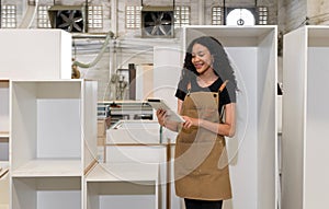 Teenager in brown apron checking email order on tablet computer. Wooden cabinets of various sizes are placed in the background.