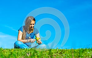 Teenager brunette girl with long hair sit on the grass and wreathes a wreath of yellow dandelion flowers on sky background with co