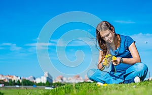 Teenager brunette girl with long hair sit on the grass and wreathes a wreath of yellow dandelion flowers on sky background with co