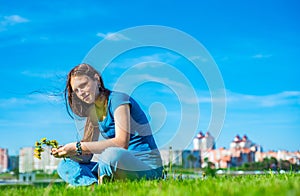 Teenager brunette girl with long hair sit on the grass and wreathes a wreath of yellow dandelion flowers on sky background with co