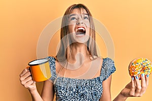 Teenager caucasian girl eating doughnut and drinking coffee angry and mad screaming frustrated and furious, shouting with anger