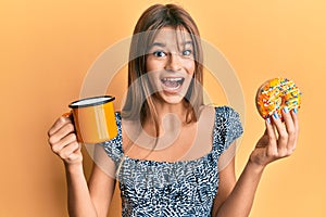 Teenager caucasian girl eating doughnut and drinking coffee celebrating crazy and amazed for success with open eyes screaming