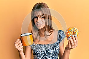 Teenager caucasian girl eating doughnut and drinking coffee clueless and confused expression