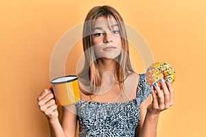 Teenager caucasian girl eating doughnut and drinking coffee relaxed with serious expression on face