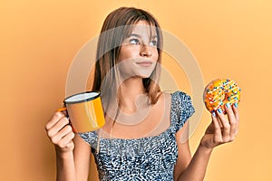 Teenager caucasian girl eating doughnut and drinking coffee smiling looking to the side and staring away thinking