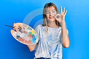 Teenager caucasian girl holding paintbrush and palette doing ok sign with fingers, smiling friendly gesturing excellent symbol