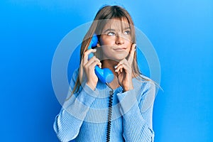 Teenager caucasian girl speaking on vintage telephone serious face thinking about question with hand on chin, thoughtful about