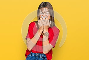 Teenager caucasian girl wearing casual red t shirt laughing and embarrassed giggle covering mouth with hands, gossip and scandal