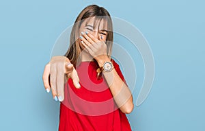 Teenager caucasian girl wearing casual red t shirt laughing at you, pointing finger to the camera with hand over mouth, shame