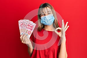 Teenager caucasian girl wearing medical mask holding 100 yuan chinese banknotes doing ok sign with fingers, smiling friendly