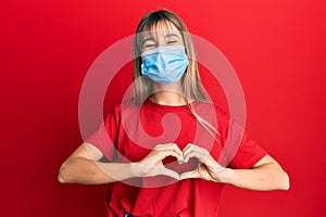 Teenager caucasian girl wearing medical mask smiling in love showing heart symbol and shape with hands