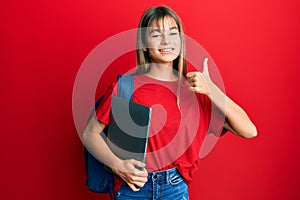 Teenager caucasian girl wearing student backpack and holding computer laptop smiling happy and positive, thumb up doing excellent