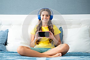 Teenager child girl showing screen phone wearing headphones listening music on smart phone sitting on bed in her room