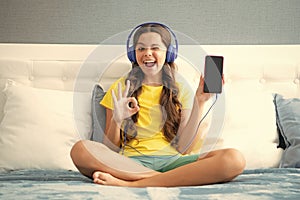 Teenager child girl showing screen phone wearing headphones listening music on smart phone sitting on bed in her room