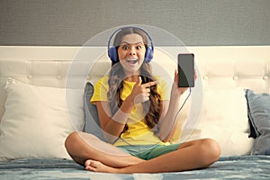 Teenager child girl wearing headphones listening music on smart phone sitting on bed in her room. Expressive emotional