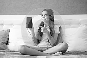 Teenager child girl wearing headphones listening music on smart phone sitting on bed in her room. Happy teen girl