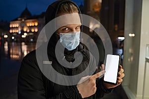 Teenager in the city wearing surgical mask pointing to smatrphone, add text.